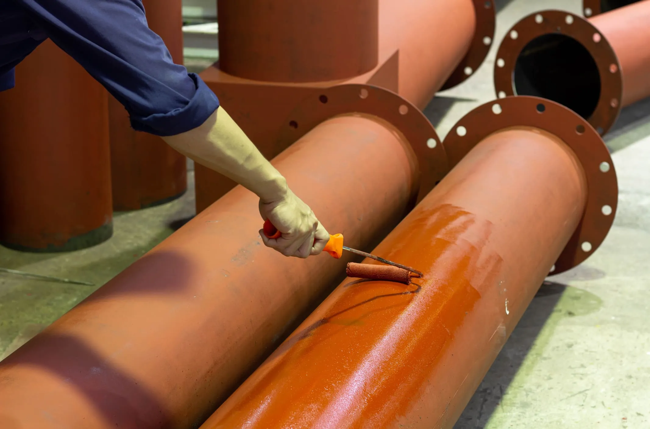 Worker rolling a protective rust-colored primer onto large flanged steel pipe sections